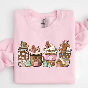 May include: A pink sweatshirt with a festive design featuring gingerbread men, candy canes, and hot chocolate. The design includes a gingerbread house, a mug of hot chocolate, and a cup of coffee.