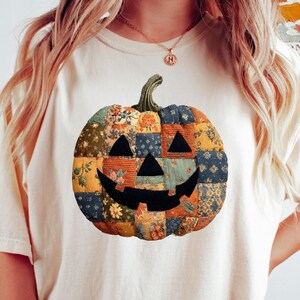 May include: Cream-colored t-shirt featuring a patchwork pumpkin design. The pumpkin has a smiling face and is made of various fabric patterns in orange, blue, and green. The shirt is a casual, fall-themed apparel item.