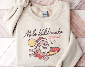May include: A beige sweatshirt with a vintage-style illustration of Santa Claus surfing. The text "Mele Kalikimaka from the land where palm trees sway" is printed on the sweatshirt.