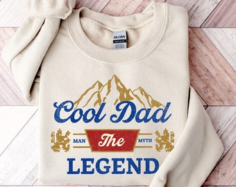 May include: A beige sweatshirt with a vintage-style graphic design that reads "Cool Dad The Legend" with a mountain range and a red banner that says "Man The Myth".