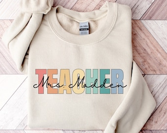 May include: A beige sweatshirt with the word "TEACHER" in rainbow colors and the text "Mrs. Madden" in black script.