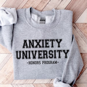 Könnte beinhalten: Grauer Pullover mit schwarzem Text, der "Anxiety University - Honors Program - " lautet.