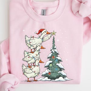 Könnte beinhalten: Rosa Sweatshirt mit einem skurrilen Weihnachtsdesign. Drei weiße Gänse sind gestapelt, mit Lichterketten und einem Weihnachtsmannhut geschmückt. Eine Gans platziert einen Stern auf einem schneebedeckten Weihnachtsbaum. Ein festliches und gemütliches Urlaubsthema.