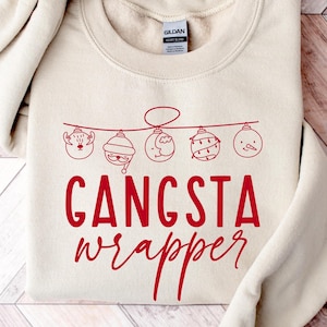 Könnte beinhalten: Cremefarbenes Sweatshirt mit dem roten Schriftzug "GANGSTA wrapper". Oberhalb des Textes befindet sich eine Reihe roter Weihnachtsschmuck-Illustrationen. Das Sweatshirt liegt gefaltet auf einer Holzoberfläche.