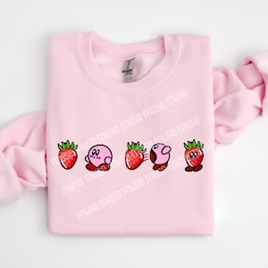 Pu&ograve; includere: Una felpa rosa chiaro con un design giocoso di un personaggio dei cartoni animati e fragole. Il design comprende cinque immagini: una fragola, un personaggio rosa, una fragola, il personaggio rosa con linee di movimento e una fragola.