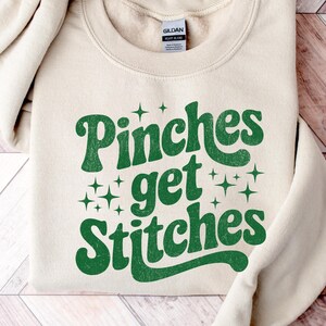 Op de afbeelding: Crèmekleurige sweatshirt met de tekst "Pinches get Stitches" in een retro groene lettertype, versierd met sterren. Het Gildan-merklabel is zichtbaar. De sweatshirt ligt op een houten oppervlak.