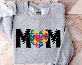 May include: Gray sweatshirt with the word "MOM" in black letters. The "O" is a heart shape made of colorful puzzle pieces with a red heart in the center.