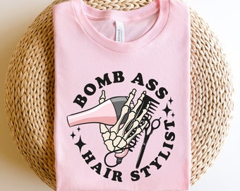 Könnte beinhalten: Pinkes T-Shirt mit einer schwarz-weißen Grafik einer Skeletthand, die einen Haartrockner und eine Schere hält. Der Text "Bomb Ass Hair Stylist" ist in einem Kreis um die Grafik geschrieben.