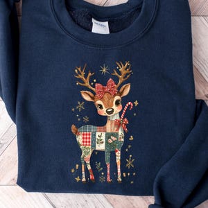 May include: Navy blue sweatshirt featuring a charming deer design. The deer has a red bow, a candy cane, and a patchwork pattern on its body. Gold snowflakes and stars surround the deer, creating a festive holiday theme.