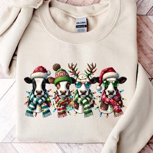 K&ouml;nnte beinhalten: Cremefarbenes Sweatshirt mit vier K&uuml;hen, die f&uuml;r Weihnachten gekleidet sind. Jede Kuh tr&auml;gt einen Weihnachtsmannhut, Rentiergeweih oder eine Winterm&uuml;tze sowie einen Schal und Lichterketten. Die K&uuml;he haben schwarz-wei&szlig;e Markierungen.