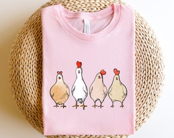Może przedstawiać: Różowy T-shirt z grafiką pięciu kurczaków w rzędzie. Kurczaki są zwrócone przodem i mają czerwone serca na głowach. Kurczaki są białe, brązowe i brzoskwiniowe.