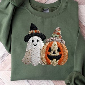 Può includere: Una felpa verde con un'applicazione a tema Halloween. Il design include un fantasma sorridente con un cappello da strega e una zucca. Il fantasma è bianco con un motivo e la zucca è arancione con una faccia nera.