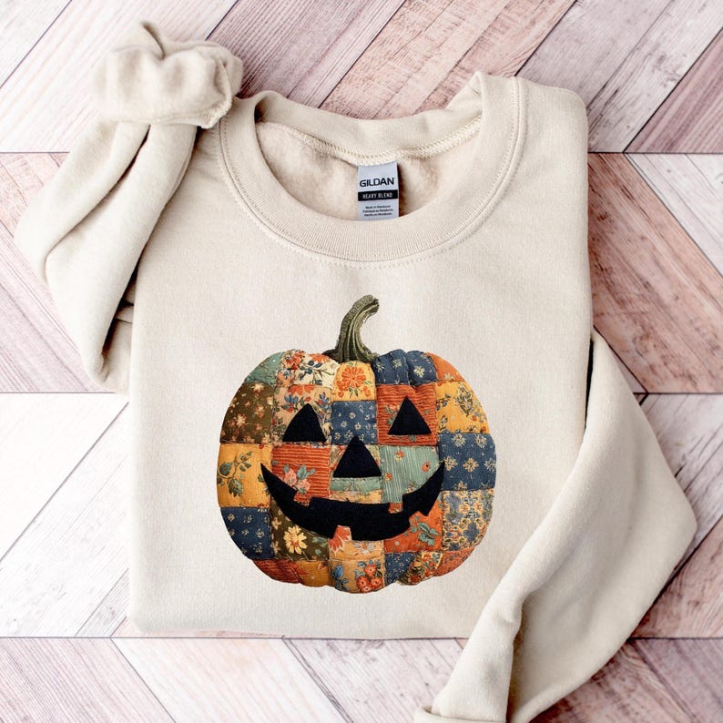 May include: A cream-colored sweatshirt featuring a patchwork pumpkin design. The pumpkin is composed of various fabric patterns in orange, blue, and floral prints, with a black face and a green stem. The sweatshirt is folded on a wooden surface.