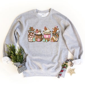 May include: Gray sweatshirt with a festive design of gingerbread men and houses on top of mugs of hot chocolate, coffee, and peppermint hot chocolate.