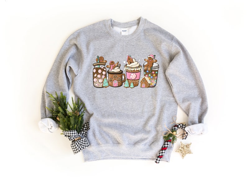 May include: Gray sweatshirt with a festive design of gingerbread men and houses on top of mugs of hot chocolate, coffee, and peppermint hot chocolate.