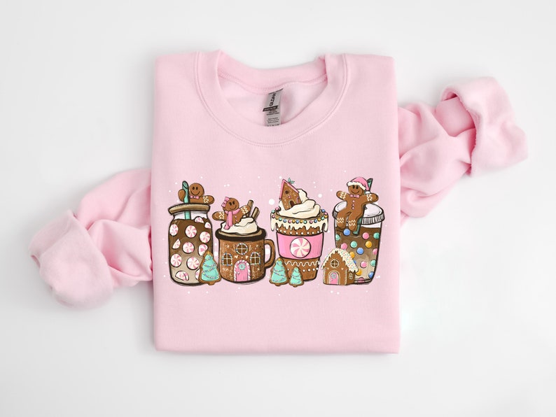 May include: A pink sweatshirt with a festive design featuring gingerbread men, candy canes, and hot chocolate. The design includes a gingerbread house, a mug of hot chocolate, and a cup of coffee.
