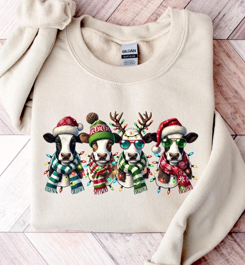 K&ouml;nnte beinhalten: Cremefarbenes Sweatshirt mit vier K&uuml;hen, die f&uuml;r Weihnachten gekleidet sind. Jede Kuh tr&auml;gt einen Weihnachtsmannhut, Rentiergeweih oder eine Winterm&uuml;tze sowie einen Schal und Lichterketten. Die K&uuml;he haben schwarz-wei&szlig;e Markierungen.