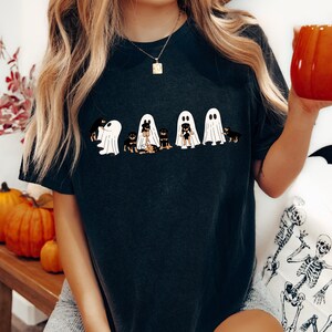 Op de afbeelding: Een zwart T-shirt met een afbeelding van vijf Rottweilers die verkleed zijn als geesten voor Halloween. De honden dragen witte geestkostuums met gezichten en staan in een rij.
