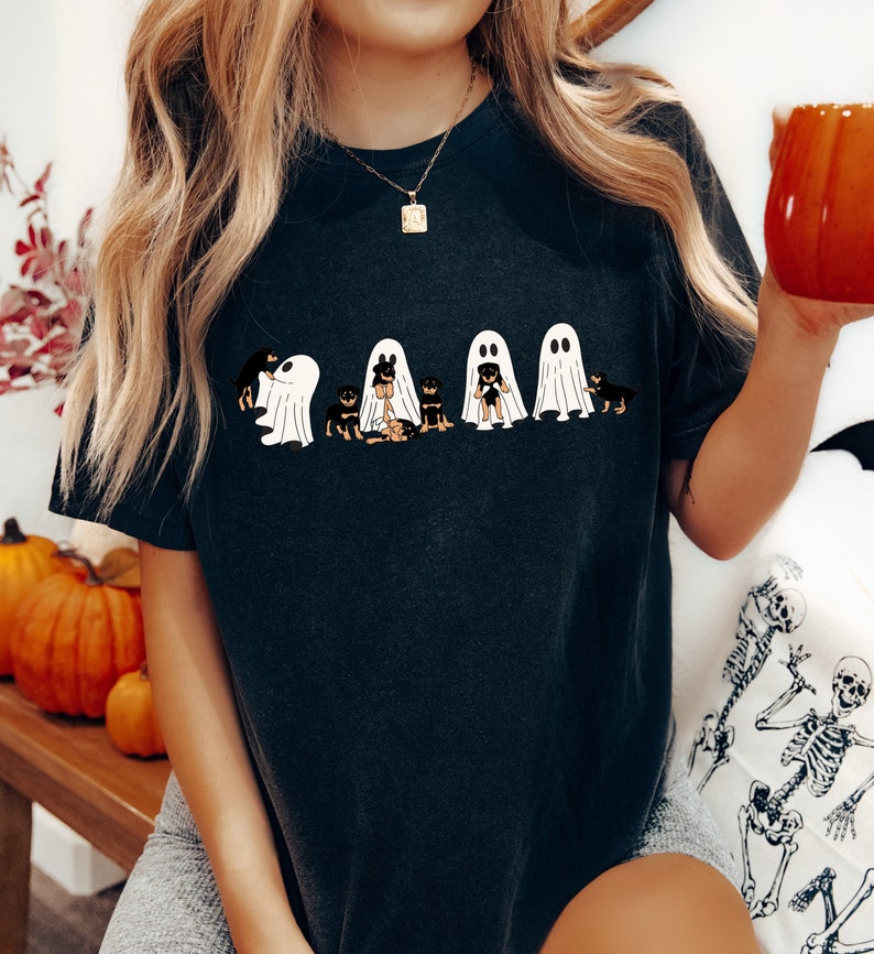 Op de afbeelding: Een zwart T-shirt met een afbeelding van vijf Rottweilers die verkleed zijn als geesten voor Halloween. De honden dragen witte geestkostuums met gezichten en staan in een rij.
