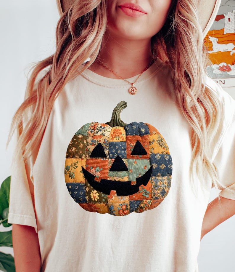 May include: Cream-colored t-shirt featuring a patchwork pumpkin design. The pumpkin has a smiling face and is made of various fabric patterns in orange, blue, and green. The shirt is a casual, fall-themed apparel item.