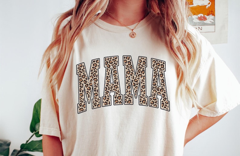 May include: A white t-shirt with the word "MAMA" printed in brown and black leopard print letters.