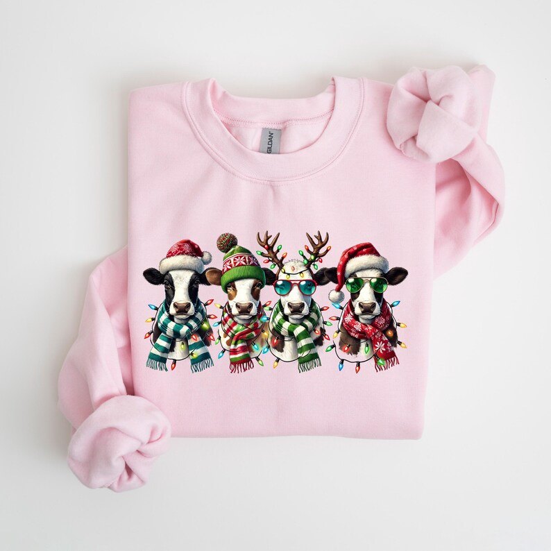 K&ouml;nnte beinhalten: Ein hellrosa Sweatshirt mit einem festlichen Design von vier Cartoon-K&uuml;hen. Jede Kuh tr&auml;gt einen Weihnachtsmannhut, Rentiergeweih oder eine Winterm&uuml;tze, eine Sonnenbrille, einen Schal und Lichterketten. Die K&uuml;he sind schwarz-wei&szlig; und braun-wei&szlig;.