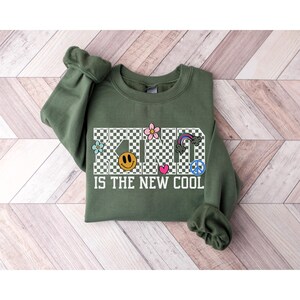 May include: Green sweatshirt with a checkered pattern that spells out "KIND IS THE NEW COOL" in white text. The word "KIND" is decorated with colorful flowers, a smiley face, a heart, a rainbow, and a peace sign.