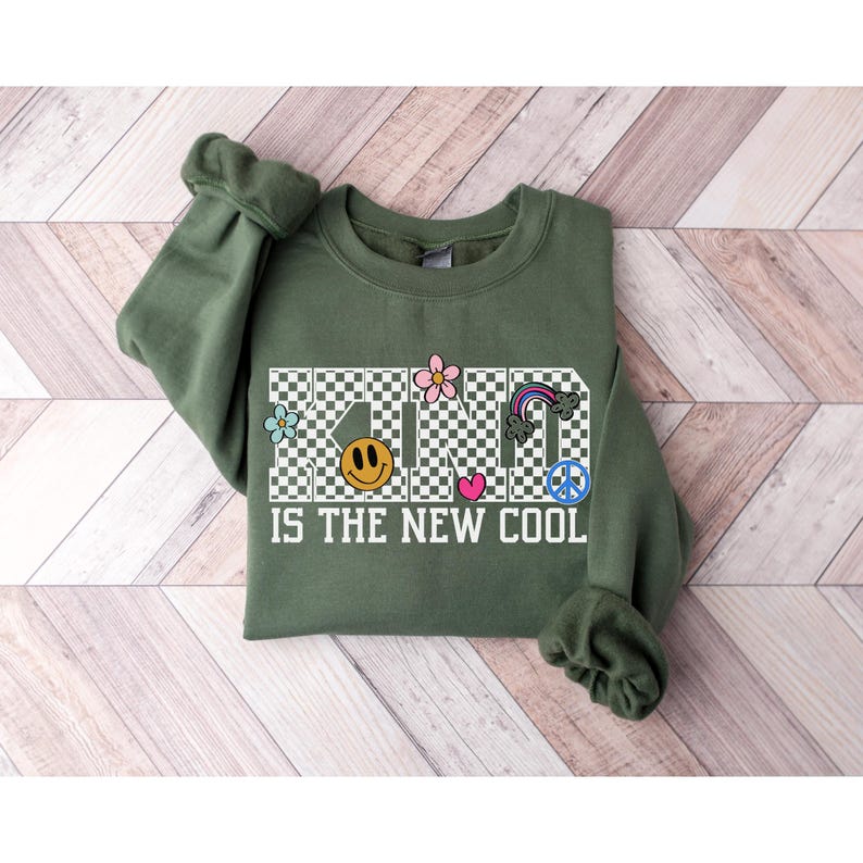 May include: Green sweatshirt with a checkered pattern that spells out "KIND IS THE NEW COOL" in white text. The word "KIND" is decorated with colorful flowers, a smiley face, a heart, a rainbow, and a peace sign.