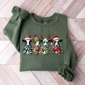 K&ouml;nnte beinhalten: Gr&uuml;nes Sweatshirt mit einem festlichen Design von vier K&uuml;hen, die jeweils mit Weihnachtsaccessoires wie Weihnachtsm&uuml;tzen, Rentiergeweihen und Sonnenbrillen geschm&uuml;ckt sind. Die K&uuml;he tragen auch Schals und sind von bunten Lichterketten umgeben.