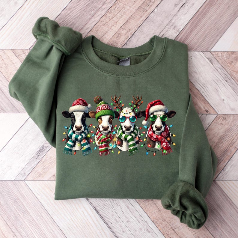 K&ouml;nnte beinhalten: Gr&uuml;nes Sweatshirt mit einem festlichen Design von vier K&uuml;hen, die jeweils mit Weihnachtsaccessoires wie Weihnachtsm&uuml;tzen, Rentiergeweihen und Sonnenbrillen geschm&uuml;ckt sind. Die K&uuml;he tragen auch Schals und sind von bunten Lichterketten umgeben.