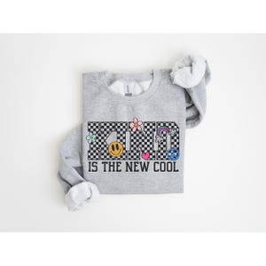 May include: Gray sweatshirt with a black and white checkered pattern that spells out "KIND" with various colorful graphic elements. The text "IS THE NEW COOL" is printed below the checkered pattern.