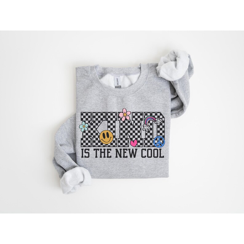 May include: Gray sweatshirt with a black and white checkered pattern that spells out "KIND" with various colorful graphic elements. The text "IS THE NEW COOL" is printed below the checkered pattern.