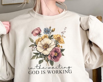 Peut inclure: Un sweat-shirt beige avec un motif floral et le texte "in the waiting GOD IS WORKING".