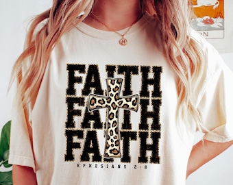 Peut inclure: Un t-shirt blanc avec le mot "FAITH" répété trois fois en noir avec une croix à imprimé léopard au centre. Le texte "EPHESIANS 2:8" est imprimé sous le motif.