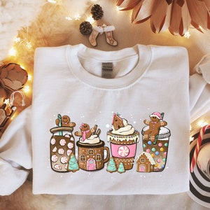 May include: White sweatshirt with a festive design featuring gingerbread men, candy canes, and hot chocolate. The design includes a gingerbread house, a cup of hot chocolate with whipped cream, and a jar of peppermint hot chocolate.
