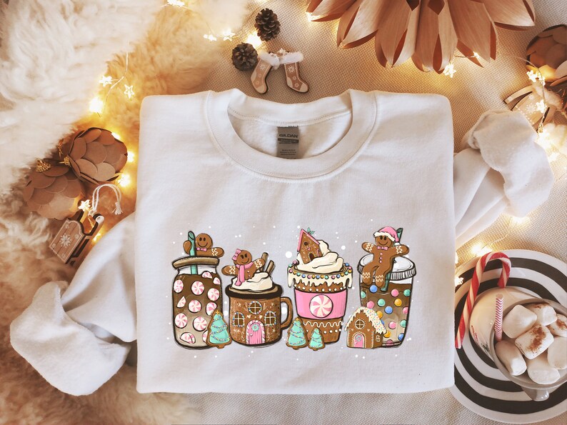 May include: White sweatshirt with a festive design featuring gingerbread men, candy canes, and hot chocolate. The design includes a gingerbread house, a cup of hot chocolate with whipped cream, and a jar of peppermint hot chocolate.