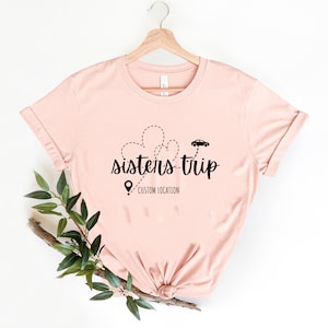 May include: A pink t-shirt with a black graphic design that says "sisters-trip" with a car and heart shapes. The text "CUSTOM LOCATION" is below the graphic.