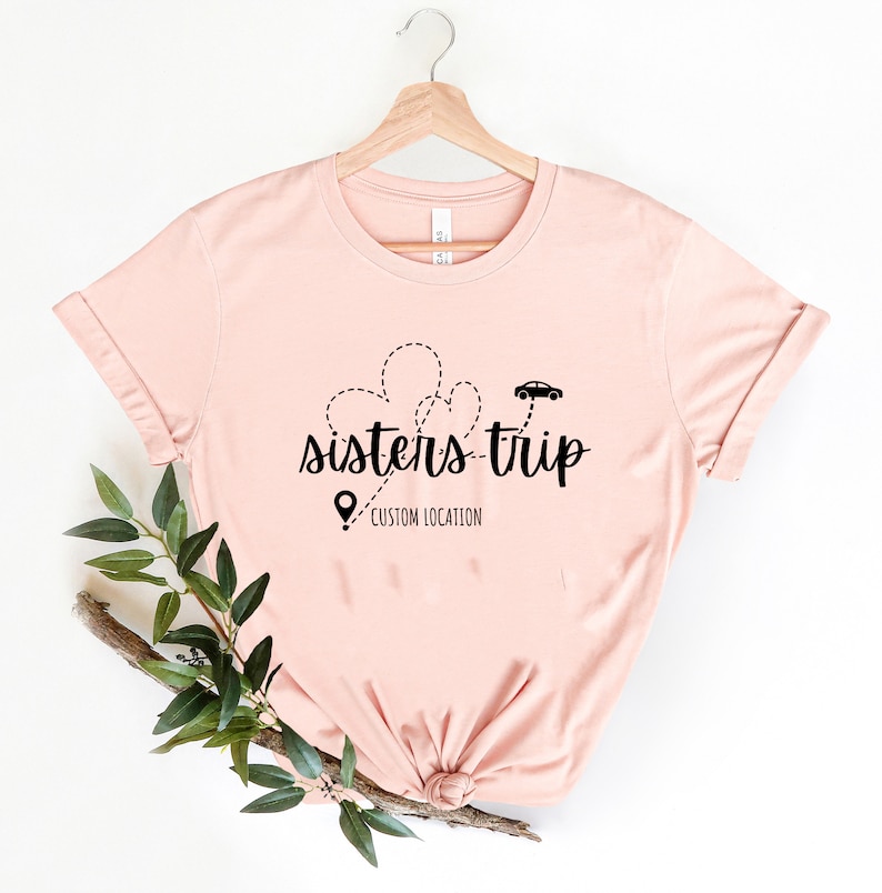 May include: A pink t-shirt with a black graphic design that says "sisters-trip" with a car and heart shapes. The text "CUSTOM LOCATION" is below the graphic.