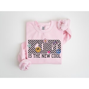 May include: Pink sweatshirt with a black and white checkered graphic that says "KIND IS THE NEW COOL" with various colorful peace signs and flowers.