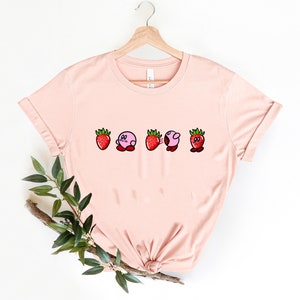 Pu&ograve; includere: T-shirt rosa chiaro con un design giocoso di una fragola e un personaggio rosa. Il design mostra il personaggio che si trasforma in una fragola. La maglietta &egrave; appesa a una gruccia di legno.