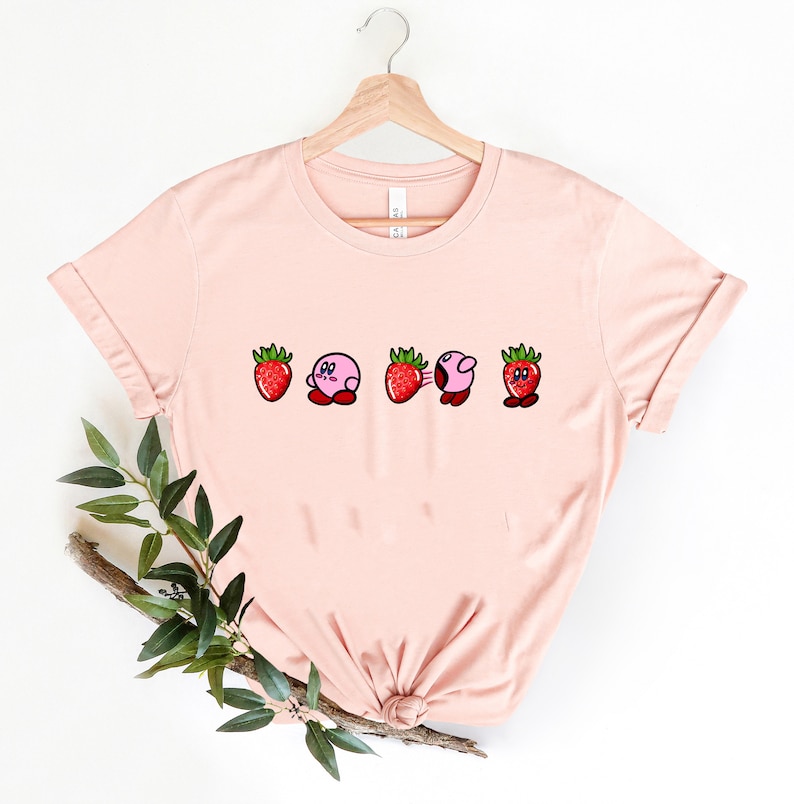Pu&ograve; includere: T-shirt rosa chiaro con un design giocoso di una fragola e un personaggio rosa. Il design mostra il personaggio che si trasforma in una fragola. La maglietta &egrave; appesa a una gruccia di legno.