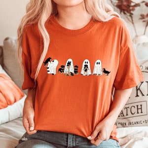 Op de afbeelding: Een feloranje T-shirt met een afbeelding van vijf zwarte en bruine honden die verkleed zijn als geesten voor Halloween. De honden dragen witte geestkostuums met zwarte ogen en monden.