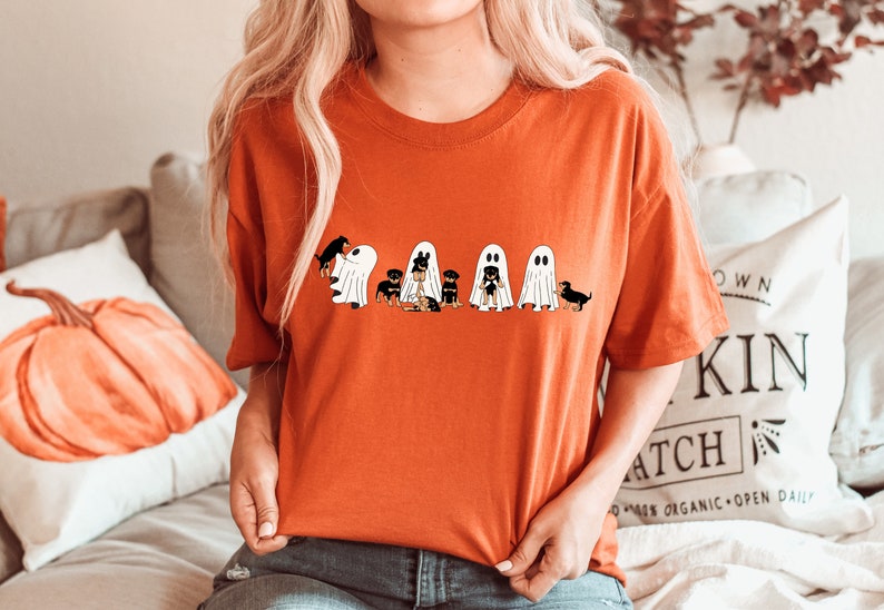 Op de afbeelding: Een feloranje T-shirt met een afbeelding van vijf zwarte en bruine honden die verkleed zijn als geesten voor Halloween. De honden dragen witte geestkostuums met zwarte ogen en monden.