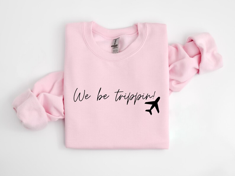 May include: Pink sweatshirt with the text "We be trippin!" and a black airplane graphic.