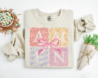 Puede incluir: Sudadera color crema con la palabra "AMEN" en cuadrados con estampado floral, acentuada por un lazo rosa. Las letras son blancas. La decoración de Pascua incluye una cesta de huevos pastel y una decoración en forma de zanahoria.