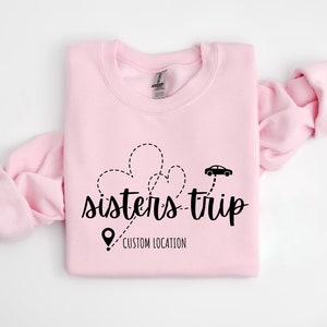 May include: Pink sweatshirt with a black graphic design that says "sisters-trip" with a car and a pin that says "custom location".