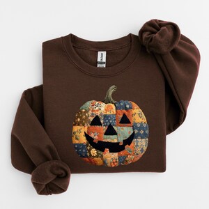 May include: A brown sweatshirt featuring a patchwork pumpkin design. The pumpkin has a smiling face and is made of various floral and patterned fabrics in shades of orange, blue, and yellow. The sweatshirt has long sleeves and a crew neck.