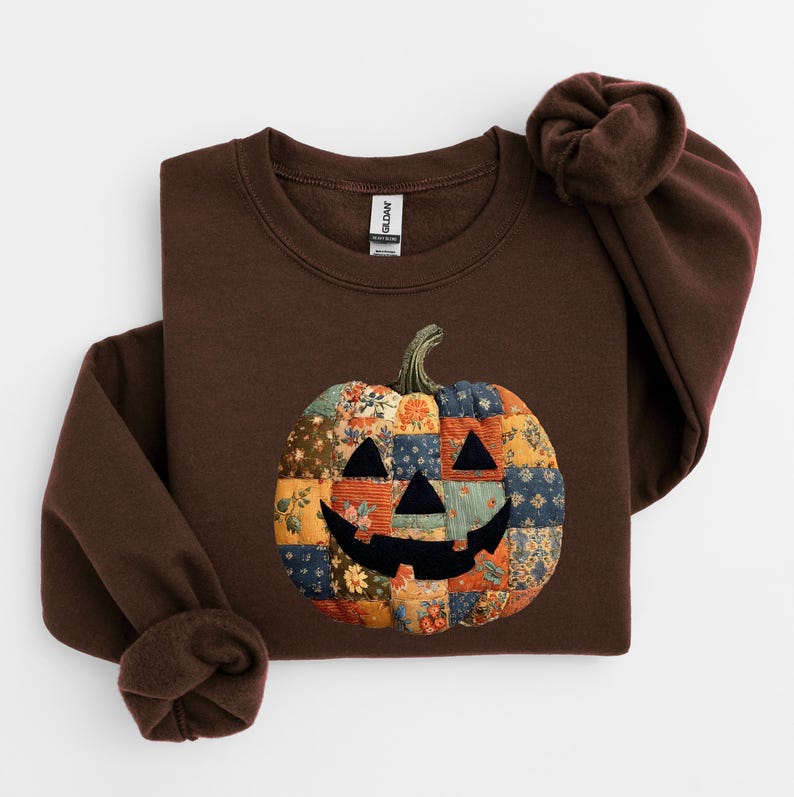 May include: A brown sweatshirt featuring a patchwork pumpkin design. The pumpkin has a smiling face and is made of various floral and patterned fabrics in shades of orange, blue, and yellow. The sweatshirt has long sleeves and a crew neck.