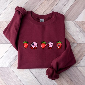 Pu&ograve; includere: Una felpa bordeaux con un design ricamato di un personaggio rosa e fragole. Il design include il personaggio in varie pose tra le fragole. Le fragole sono rosse con foglie verdi. La felpa &egrave; realizzata in materiale morbido.