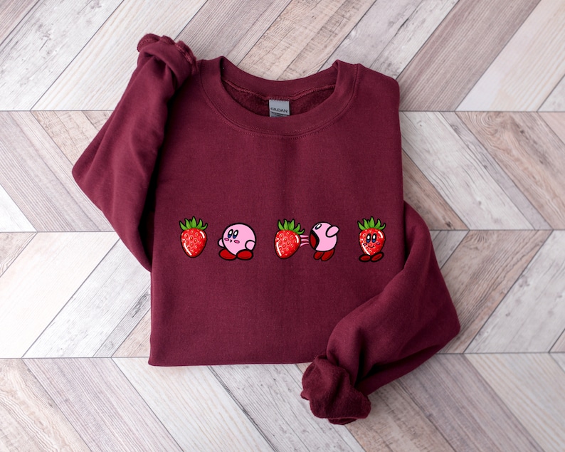 Pu&ograve; includere: Una felpa bordeaux con un design ricamato di un personaggio rosa e fragole. Il design include il personaggio in varie pose tra le fragole. Le fragole sono rosse con foglie verdi. La felpa &egrave; realizzata in materiale morbido.